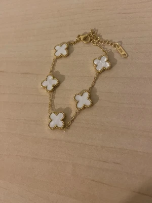 Guldfärgat armband med vita blommor - Säljer ett elegant armband i guldfärg med fem vita fyrklöverformade hängen. Kedjan är tunn och har ett justerbart lås. Hängena har en pärlemorskimrande yta och är inramade av små guldfärgade kulor. Perfekt accessoar för att lyfta din stil.