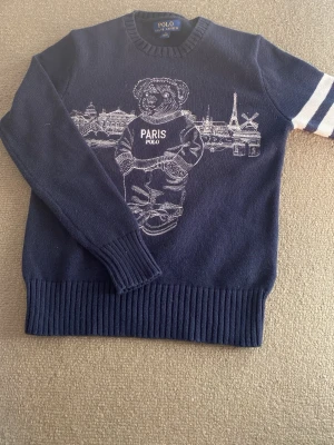 Mörkblå stickad tröja Polo Bear - Mörkblå stickad tröja från Polo Ralph Lauren med Polo Bear-motiv och Paris-tryck på bröstet. Tröjan har rund hals, långa ärmar och två vita ränder på ena ärmen. Ribbstickade muddar vid ärmslut och nederkant.
