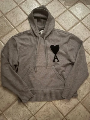 Ami Paris tröja  - Snygg grå hoodie från Ami Paris med svart hjärta och bokstaven A broderat på bröstet. Hoodien har huva,magficka och ribbade muddar. Perfekt för en chill och trendig look. Storlek S,nästan helt oanvänd! Hör bara av er om ni undrar något!