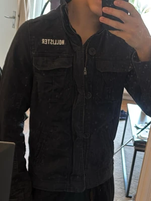 Svart jacka från Hollister med dragkedja - Säljer en svart jacka från Hollister med dragkedja och knappar framtill. Jackan har två bröstfickor med lock och två sidofickor. Klassisk krage och broderad Hollister-logga på bröstet. Perfekt för dig som gillar en stilren och enkel look.