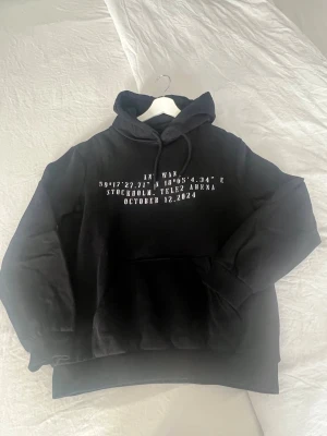 Svart hoodie ANTWAN Tele2 Arena - Säljer en svart hoodie från ANTWAN med tryck fram och bak. Framsidan har vit text med koordinater och datum för Tele2 Arena, Stockholm. Baksidan har en stor färgglad grafik med texten ANTWAN och motiv. Hoodien har huva med dragsko och magficka.