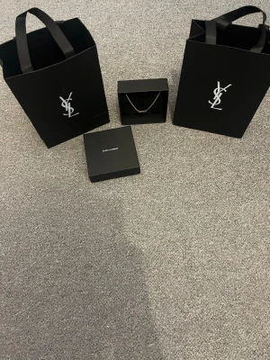 Guldfärgat armband från Yves Saint Laurent - Snyggt guldfärgat armband från Saint Laurent med den ikoniska YSL-loggan som hänge. Kedjan är tunn och elegant, perfekt för att lyfta vilken outfit som helst. Levereras i originalask och med exklusiva Saint Laurent-påsar.