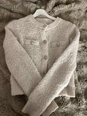 Beige stickad kofta med knappar - Mysig beige stickad kofta med rund halsringning och dekorativa knappar framtill. Koftan har två fejkfickor med knappar på bröstet och ribbade muddar vid ärmslut och nederkant. Perfekt för lager-på-lager-stil.