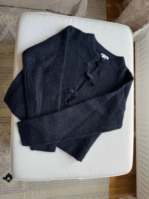 Mörkgrå stickad tröja med rosetter - Mysig mörkgrå stickad tröja med rund halsringning och dekorativa svarta rosetter framtill. Tröjan har lång ärm och en enkel, rak passform. Perfekt för dig som gillar en stilren look med en söt detalj.