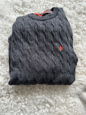 Mörkgrå kabelstickad tröja Morris - Mörkgrå kabelstickad tröja från Morris med rund halsringning och röd broderad logga på bröstet. Tröjan har ribbade muddar och är tillverkad i mjukt material, perfekt för lager-på-lager-stil.