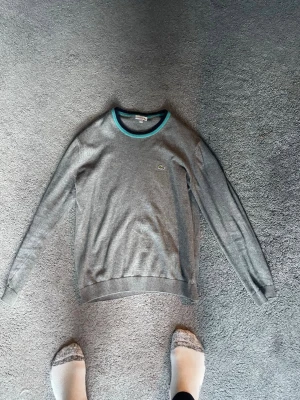 Grå tröja från Lacoste - Snygg grå tröja från Lacoste med rund halsringning som har blå och turkosa detaljer. Liten Lacoste-logga på bröstet. Perfekt för dig som gillar klassisk och stilren design.