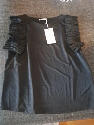 Svart topp med volangärmar från Lindex - Trendig svart topp från Lindex med unika volangdetaljer på axlarna. Toppen har rund halsringning och är ärmlös med breda axelpartier som ger en cool siluett. Perfekt för dig som vill sticka ut lite extra.