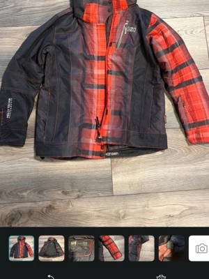 Röd och svart vindjacka Helly Hansen - Snygg vindjacka från Helly Hansen i rött och svart rutigt mönster. Jackan har hög krage, dragkedja, flera fickor och justerbara ärmslut. Perfekt för blåsiga dagar och har en sportig look med logga på bröstet. Materialet är slitstarkt och vattenavvisande.