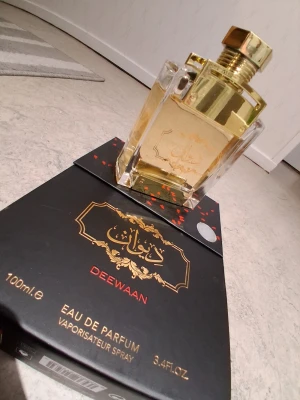 Deewaan Eau de Parfum 100ml - Lyxig parfymflaska från Deewaan med guldfärgad detalj och genomskinligt glas. Flaskan har en elegant, fyrkantig form och arabiska inskriptioner. Kommer i en svart och guld kartong med exklusiv design. Innehåller 100 ml Eau de Parfum.