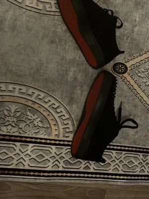 Svarta Louboutin sneakers med nitar - Säljer ett par svarta sneakers från Christian Louboutin med klassiska röda sulor och coola nitar på tån. Skorna har snörning och är tillverkade i mocka och läder. Perfekta för dig som vill sticka ut med en edgy och lyxig stil. Använd bara en gång 