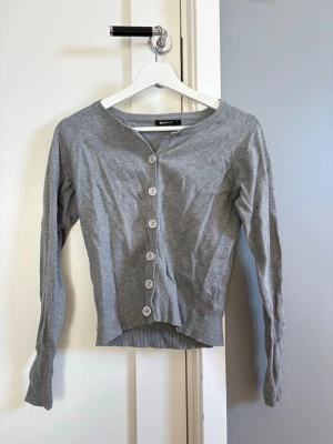 Grå kofta från Gina Tricot - Snygg grå kofta från Gina Tricot med rund halsringning och långa ärmar. Koftan har ribbade muddar vid ärmslut och nederkant samt knäppning med sex matchande knappar framtill. 