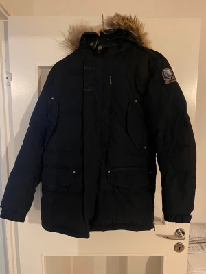 Svart Parajumpers parkas med päls - Svart parkas från Parajumpers med huva och avtagbar päls. Jackan har flera fickor framtill, dragkedja och tryckknappar. Klassisk patch på ärmen och varm vaddering, perfekt för kalla dagar.