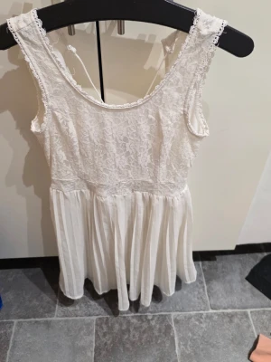 Vit spetsklänning med volang - Söt vit klänning med spets upptill och volangkjol. Klänningen är ärmlös och har rund halsringning samt knyt i ryggen. Perfekt för sommarens alla tillfällen och ger en romantisk känsla med sitt tunna, luftiga tyg.