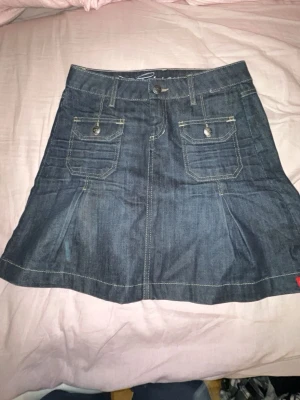 Mörkblå jeanskjol med fickor - Säljer en mörkblå jeanskjol från esprit med två stora fickor framtill och baktill, dekorativa sömmar och knappstängning i midjan. Kjolen har en klassisk A-linjeform och detaljer i silverfärgade knappar. Perfekt för dig som gillar denimstil.