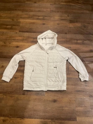 Vit cardigan jacka - Stilren vit vindjacka med quiltat mönster och dragkedja framtill. Jackan har två fickor med dragkedja och en praktisk huva. Perfekt för blåsiga dagar och enkel att matcha med olika outfits. Tillverkad i lätt material för extra komfort.