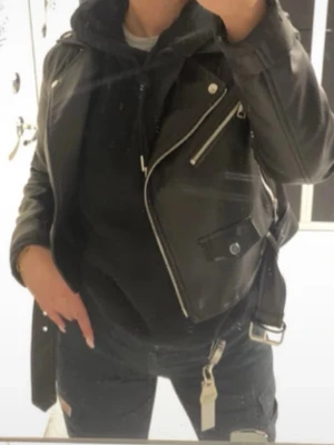 Svart biker skinnjacka med dragkedjor - Snygg svart skinnjacka i klassisk bikerstil med silvriga dragkedjor och knappar. Jackan har breda slag, snedställd dragkedja och detaljerade fickor. Perfekt att slänga över en hoodie eller skjorta för en cool look.