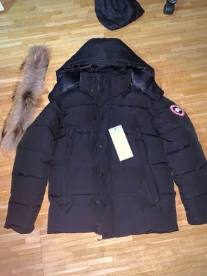  Canada Goose puffer  - Snygg svart dunjacka från Canada Goose med avtagbar huva och pälskant. Jackan har klassisk patch på ärmen, dragkedja och knappar framtill samt flera fickor. Perfekt för kalla vinterdagar och riktigt varm tack vare dunfyllningen.