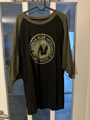Svart/grön Gorilla Wear t-shirt - Cool svart t-shirt från Gorilla Wear med kontrasterande mörkgröna ärmar och tryck fram och bak. Stort logotryck på bröstet och hashtag på sidan. Perfekt för dig som gillar streetwear och sportig stil.