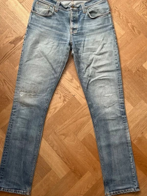 Ljusblå slitna jeans från Nudie - Säljer ett par ljusblå jeans från Nudie med raka ben och slitningar på både fram- och baksida. Klassisk femficksmodell med snygga kontrastsömmar och broderade bakfickor. Tillverkade i denim av bomull. Perfekta för en avslappnad look.