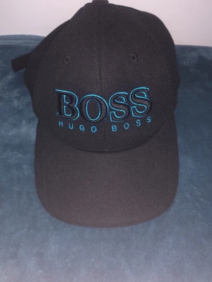 Svart keps från Hugo Boss - Snygg svart keps från Hugo Boss med broderad blå logga framtill. Klassisk böjd skärm och justerbar rem baktill. Kepsen är tillverkad i ett strukturerat tyg som ger en cool och sportig känsla. Perfekt accessoar för en stilren streetlook.
