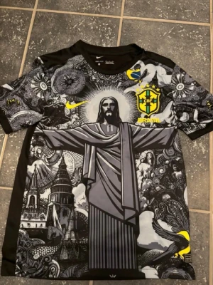 Nike Brazil Jesus grafisk fotbollströja - Unik svart och grå Brasilien-tröja från Nike med stort grafiskt tryck av Kristusstatyn, detaljerade illustrationer och gula accenter. CBF-logga och Nike Swoosh på bröstet. Tillverkad i lätt och ventilerande material, perfekt för fotboll eller streetwear. Splitter ny!👍