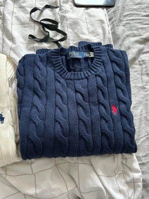 Mörkblå kabelstickad tröja Polo Ralph Lauren - Mörkblå kabelstickad tröja från Polo Ralph Lauren med rund halsringning och klassisk röd logga broderad på bröstet. Tröjan har långa ärmar och ribbade muddar. Perfekt för en stilren och tidlös look.
