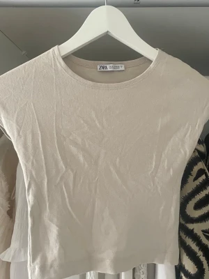 Beige croppad topp från Zara - Snygg croppad topp från Zara i beige färg. T-shirten har rund halsringning och korta ärmar. Tillverkad i mjukt bomullsmaterial som sitter tight och bekvämt. Perfekt att matcha med höga jeans eller kjol.
