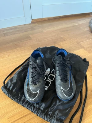 Nike Mercurial svarta fotbollsskor - Snygga svarta Nike Mercurial fotbollsskor med blå detaljer och vit swoosh. Skorna har snörning och en strömlinjeformad design för snabbhet på planen. Ovandelen är i syntetmaterial och innersulan är blå. Perfekta för dig som vill sticka ut på matchen. Lite trasig vid snörningen på höger Fot. Påse ingår