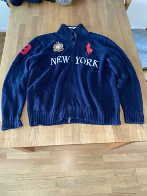 Marinblå ziptröja från Ralph Lauren - Snygg marinblå ziptröja från Ralph Lauren med broderad röd logga, texten NEW YORK på bröstet och patchdetaljer på ärmen. Tröjan har hög krage, ribbade muddar och är tillverkad i ett mjukt stickat material. Perfekt för dig som gillar sportig och klassisk stil.