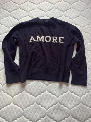 Svart stickad tröja AMORE Gina Tricot - Cool marinbkå tröja från Gina Tricot med vit text 'AMORE' framtill. Tröjan har rund halsringning och långa ärmar, perfekt för en avslappnad och trendig look. Den är lite nopprig men syns ej så mycket.