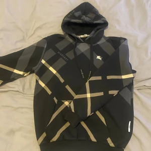Burberry svart hoodie med rutmönster - Snygg svart hoodie från Burberry med klassiskt rutmönster i beige och grått. Stor huva med dragsko och broderad Burberry-logga på bröstet. Tillverkad i mjukt bomullsmaterial som känns riktigt skönt mot huden. Perfekt för dig som vill ha en stilren och lyxig streetwear-look. Bra skick, pris går atg diskutera. Storlek L men passar M-L