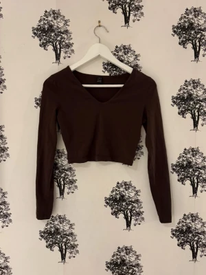 Brun croppad långärmad topp - Mörkbrun croppad topp med lång ärm och v-ringning. Enkel och stilren design som passar perfekt till jeans eller kjol. Tillverkad i mjukt bomullsmaterial för en bekväm känsla.