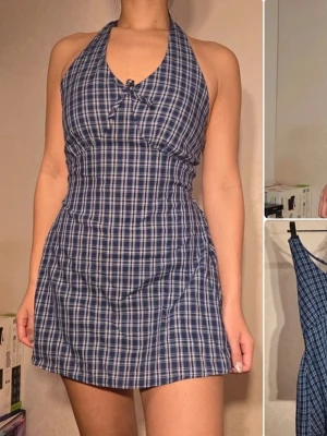 Blå rutig kortklänning från EGGKA - Supersöt blå och vit rutig kortklänning från EGOKA med halterneck och liten rosett framtill. Klänningen har figurnära passform och är ärmlös, perfekt för varma dagar. Tillverkad i bomull för en skön känsla.