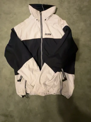 Peak Performance svartvit vindjacka - Snygg svartvit vindjacka från Peak Performance med hög krage och dragkedja framtill. Jackan har två fickor med dragkedja, justerbar nederkant och logga på bröstet samt baktill. Perfekt för blåsiga dagar och sportiga outfits.