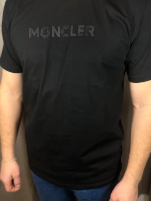 Svart Moncler t-shirt i bomull - Snygg svart t-shirt från Moncler med diskret logotyp i svart och grått på bröstet. Klassisk passform och rund halsringning. Tillverkad i mjuk bomull som känns skön mot huden. Perfekt till jeans eller shorts.