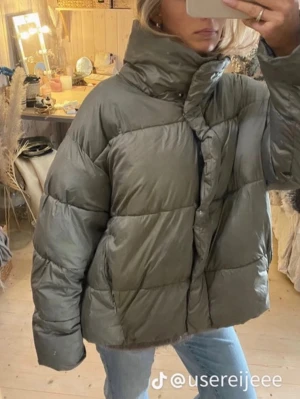 Olivgrön pufferjacka med hög krage - Säljer en olivgrön pufferjacka från H&M med oversized passform och hög krage. Jackan har dragkedja och tryckknappar framtill samt två sidofickor. Perfekt för kalla dagar och riktigt skön att bära. Materialet är syntet och jackan är quiltad för extra värme.