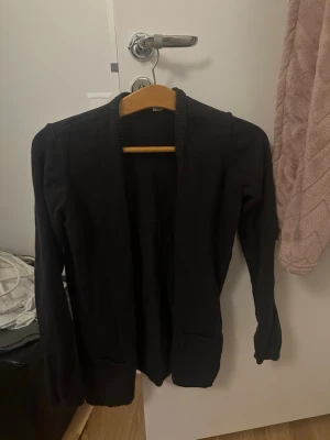 Svart stickad kofta med fickor - Svart stickad kofta med lång ärm och öppet framstycke. Koftan har två praktiska fickor framtill och ribbad kant runt halsen. Perfekt att slänga på sig när det är lite kyligt ute.