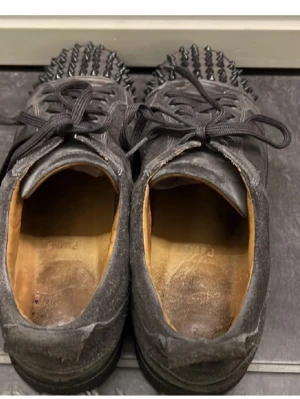 Gråa loubs med nitar  - Unika svarta sneakers med rund tå och platt sula, täckta med svarta nitar på tån. Skorna har snörning och är gjorda i mocka med läderfoder på insidan. Perfekt för dig som vill sticka ut med en edgy stil och gillar detaljer som syns.