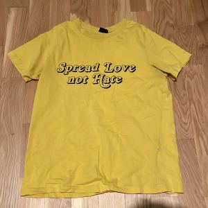 Gul t-shirt med tryck från Gina Tricot - Gul t-shirt från Gina Tricot i storlek XS med svart texttryck 'Spread Love not Hate' på framsidan. Klassisk rund hals och korta ärmar, tillverkad i mjuk bomull. Perfekt för dig som vill ha en färgglad och statement-plagg i garderoben.