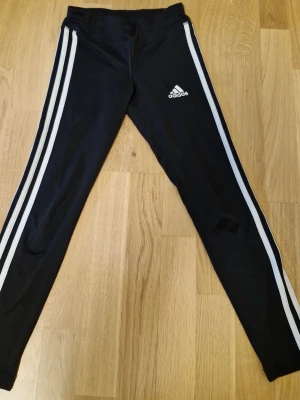 (Barnkläder) Svarta Adidas leggings med vita ränder - Säljer ett par svarta Adidas leggings med klassiska vita ränder längs sidorna och Adidas-logga på låret. Storlek 140 cm. De är tillverkade i stretchigt material med 91% polyester och 9% elastan, vilket gör dem riktigt sköna och följsamma. Perfekta för träning eller chill.
