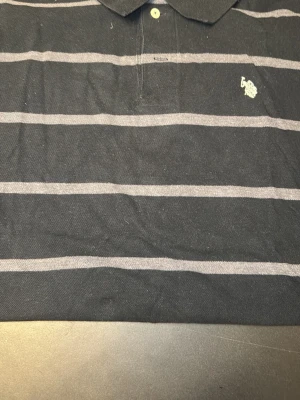 Svart och grå randig polotopp - Säljer en svart polotopp från Polo Ralph Lauren med gråa horisontella ränder och klassisk krage. Toppen har en broderad logga på bröstet och knappar vid halsen. Perfekt för dig som gillar stilren design med sportig touch.