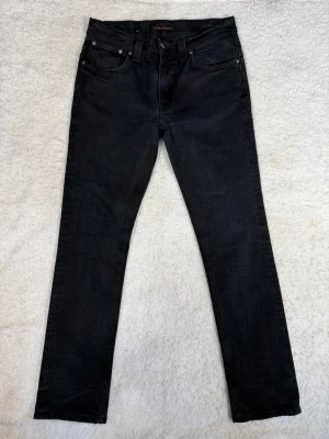  Nudie Jeans - storlek W32/L32 - Snygga svarta jeans från Nudie Jeans med klassisk femficksdesign och silverfärgade nitar. Modellen är skinny med smal passform och raka ben. Jeansen har normal midja och är tillverkade i mjukt bomullsmaterial med stretch för extra komfort.