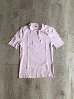 Vintage topp med knyt - Supersöt ljusrosa topp från Anne Weyburn med korta ärmar och knyt i halsen. Toppen har ribbade muddar vid ärmslut och nederkant. Toppen är gjord av syntet och har en feminin och klassisk stil. Observera en röd färgfläck på nedre mudden. 