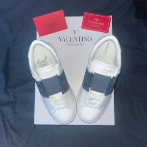 Valentino Garavani Open sneakers vit/blå - Säljer ett par Valentino Garavani Open sneakers i vitt skinn med bred mörkblå rem över snörningen. Snygga detaljer med guldlogga på plösen och innersulan samt mönstrad vit yttersula. Perfekt för dig som gillar lyxiga och stilrena sneakers. (Säljs pga att dom inte passar, köptes på Plick)
