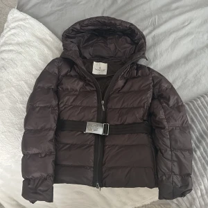 Mörkbrun pufferjacka från Moncler - Snygg mörkbrun pufferjacka från Moncler med huva och ett brett bälte med silverfärgat spänne med loggan. Såklart äkta, kan fixa äkthets bevis för intresserade. Strl 2 står inuti, gammal modell.  Päls hör till skriv vid intresse och fler bilder med den också!