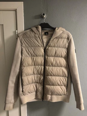 Beige pufferjacka med huva från Duno - Snygg beige jacka  från Duno. Jackan har huva, dragkedja och svart logotyp på ärmen. Köpt på Joel i borås nypris cirka 4000. Pris kan diskuteras. 