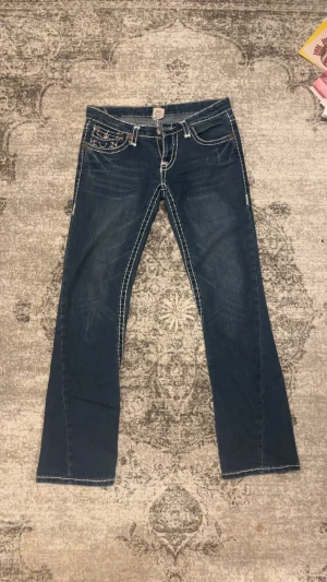 True Religion Joey Super T jeans, blå, W30 - Säljer ett par True Religion Joey Super T jeans( low waist)  i klassisk blå denim med kontrasterande vit söm och snygga detaljer på bakfickorna. Passar även W29 . Jeansen har bootcut-modell/Straight och är tillverkade i jeansmaterial. Märkta med True Religion-logga och tillverkade i USA. (Köpte den för över 1800kr ) 
