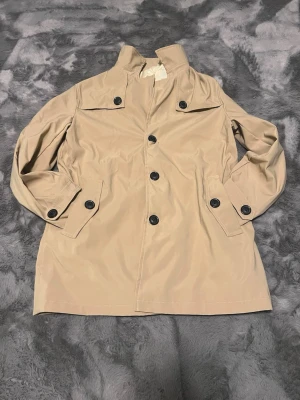 Beige trenchcoat med svarta knappar - Snygg beige trenchcoat med klassisk krage och svarta knappar framtill. Jackan har långa ärmar med knappdetaljer vid mudden och två fickor. Insidan är fodrad i ljus satinliknande material. Perfekt för dig som gillar stilrena och tidlösa plagg. Ingen tecken på användning. Inte i orginal förpackning.