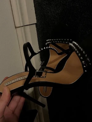 Svarta sandaler med nitar från Zara - Säljer ett par svarta sandaler från Zara med smala remmar och coola silverfärgade nitar längs ovansidan. Skorna har en smal hög klack och spänne vid ankeln. Perfekt för dig som gillar edgy detaljer och stilren design. De är i storlek 40 men sitter perfekt på mig som bär 38-39❤️