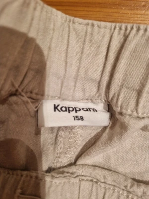Beige linnebyxor från Kappahl, strl 158 - Snygga och luftiga beige linnebyxor från Kappahl i storlek 158. Byxorna har rak passform, resår i midjan och snörning för extra komfort. Perfekta för varma dagar och chill stil. Fickor på sidorna för det viktigaste.